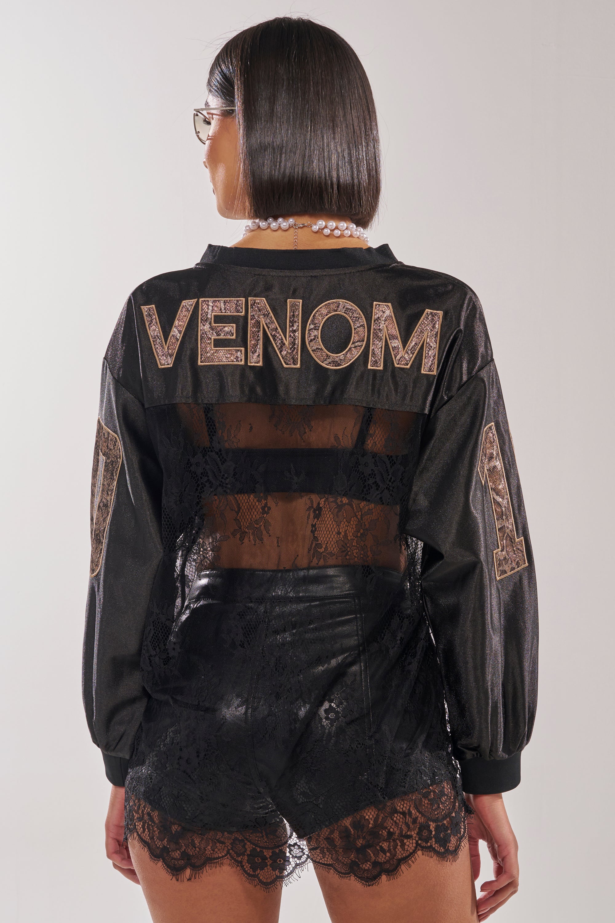 A woman with a sleek bob stands with her back to the camera, streetwear ready in an oversized ROCKY LACE JERSEY and a black jacket featuring "VENOM" and "1" in varsity-style lettering against a plain background. 5