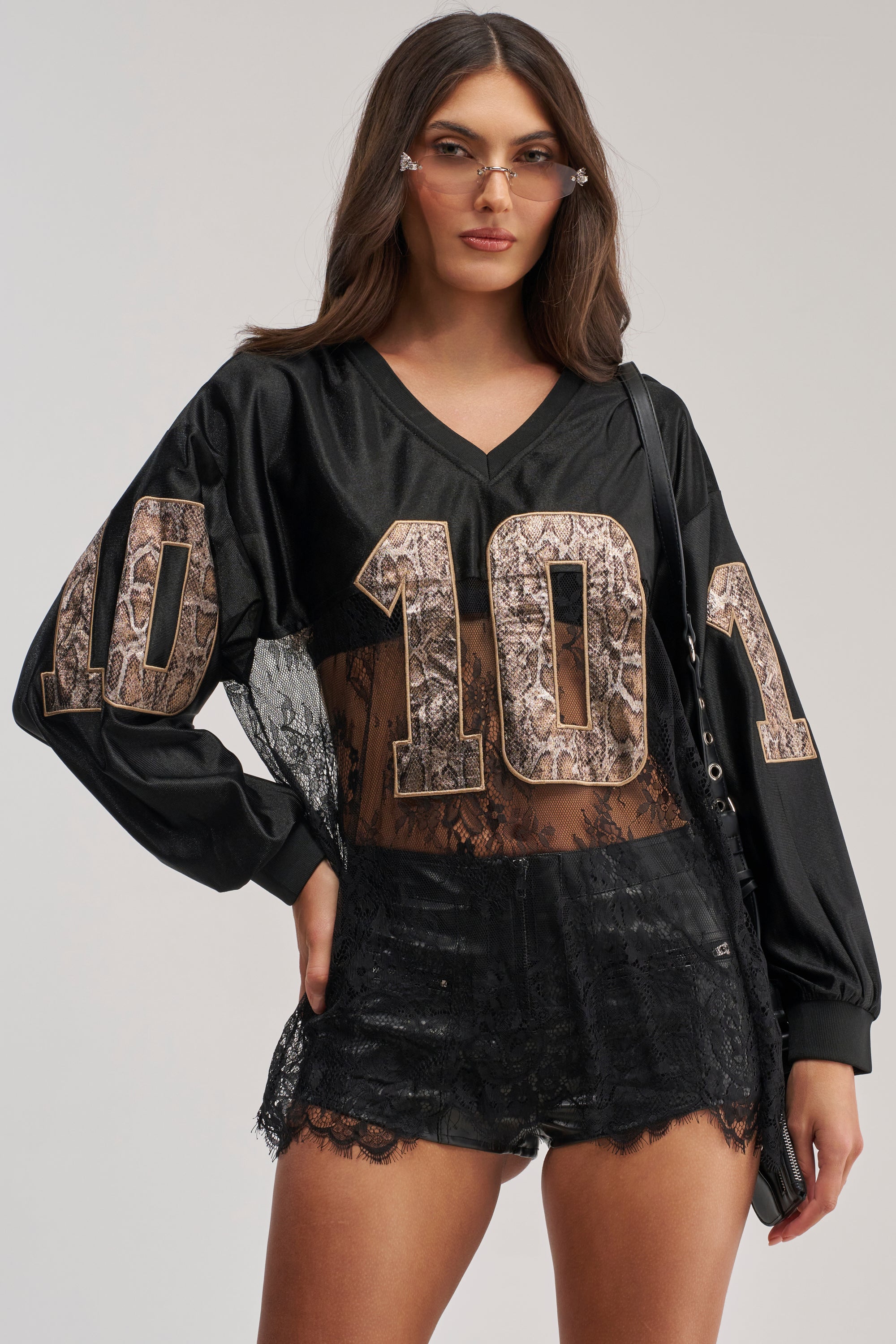 A woman models the ROCKY LACE JERSEY—a black varsity-style top with bold snake-print "101"—paired with black lace shorts, clear glasses, and a black shoulder bag, highlighting her unique streetwear look against a simple backdrop.