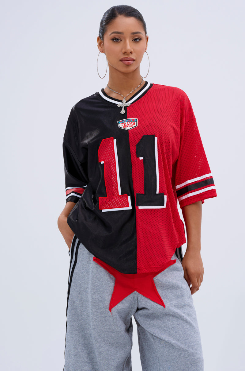 A woman models the SPLIT PERSONALITY JERSEY TOP, featuring a half black, half red color block design with “11” on the front. She pairs it with gray pants with a red star at the waist, large hoop earrings, and pulled-back hair.