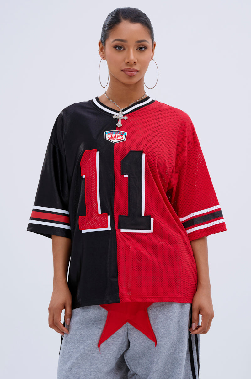 A woman faces forward against a plain light background, wearing the SPLIT PERSONALITY JERSEY TOP—half black, half red with number 11—paired with large hoop earrings and grey pants featuring a red star.