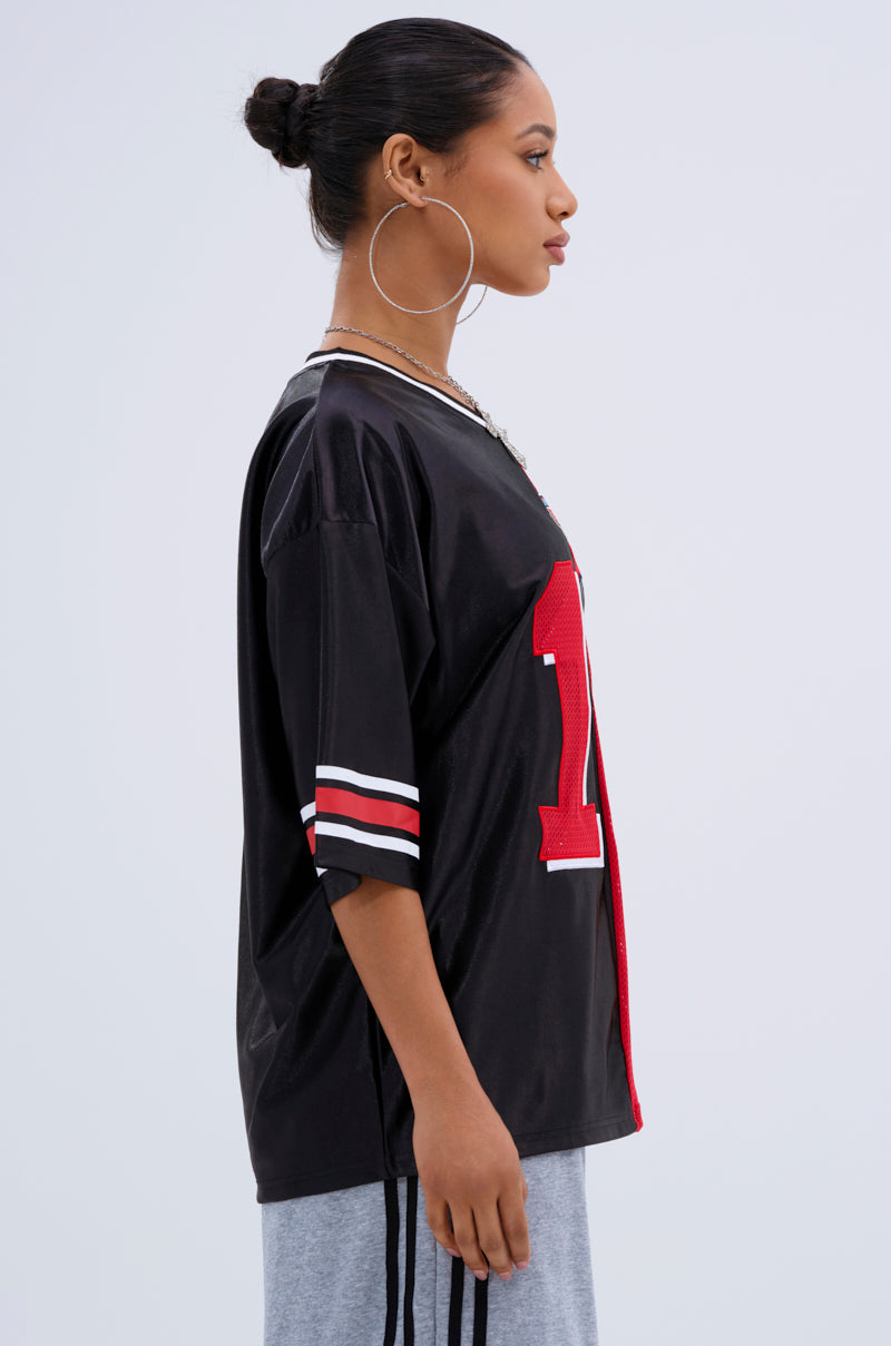 A woman with her hair in a bun and large hoop earrings wears the SPLIT PERSONALITY JERSEY TOP, featuring a color block red and white stripe design with the number 1, paired with gray pants, as she stands in profile against a light background.