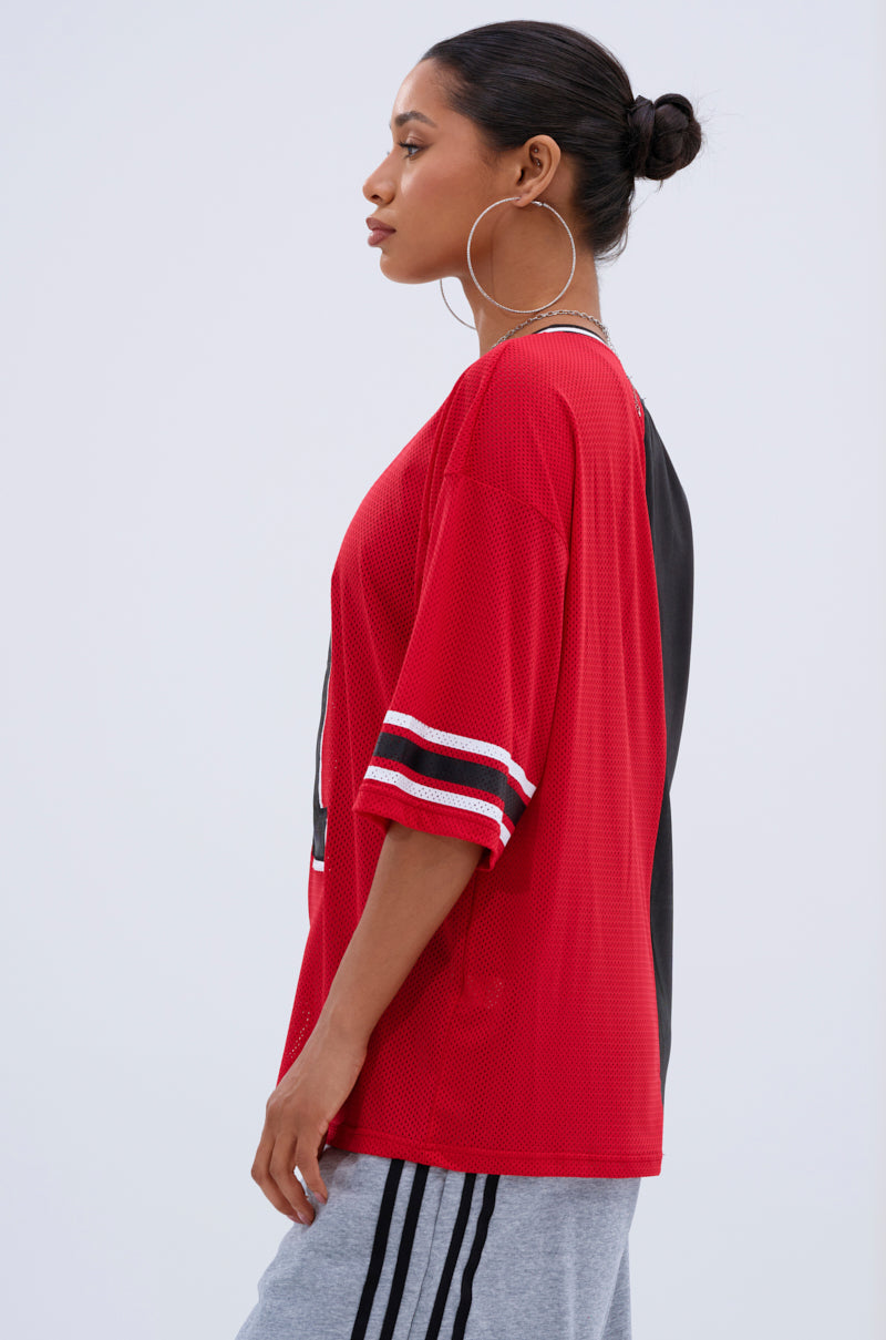 A woman in profile stands against a light background wearing the SPLIT PERSONALITY JERSEY TOP with white and red striped sleeves, gray sweatpants with black stripes, large hoop earrings, and her hair tied back.