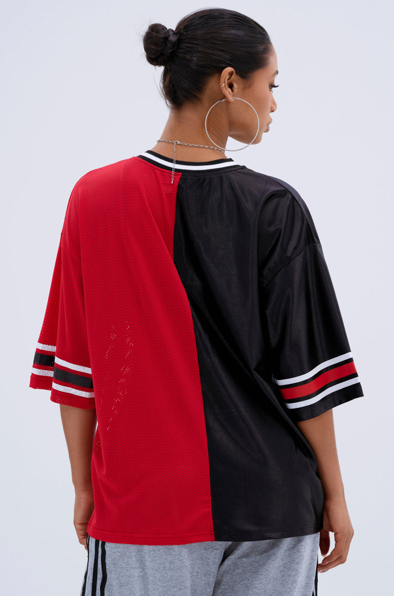 A person with dark hair in a bun and large hoop earrings wears the SPLIT PERSONALITY JERSEY TOP featuring a bold red and black color block design with striped cuffs, standing with their back to the camera.