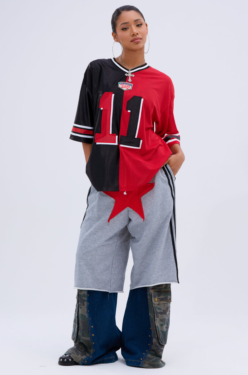 A woman stands confidently in the SPLIT PERSONALITY JERSEY TOP with bold color blocking, paired with loose gray star-print shorts and layered blue camo pants. She completes the look with hoop earrings and a tied-back hairstyle.