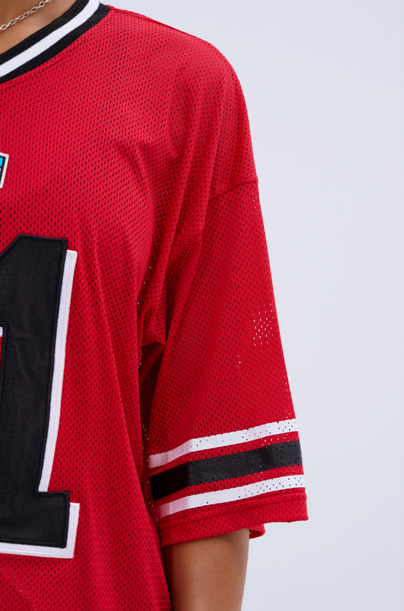 Someone wears the SPLIT PERSONALITY JERSEY TOP, showcasing a red color block design, white and black sleeve stripes, and a large black number on the front. The background is plain and light-colored.