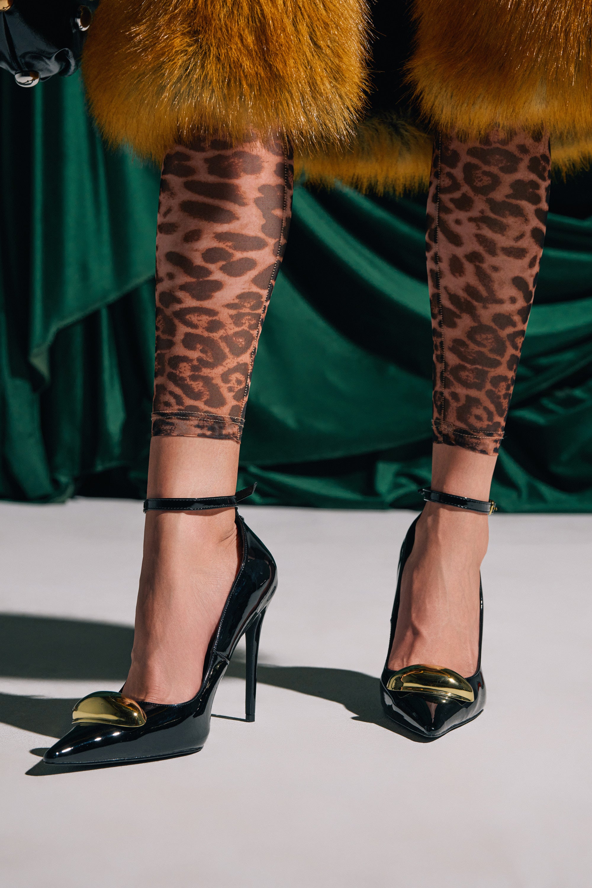 A person in AZALEA WANG ESMARIA BLACK STILETTO PUMPS, sheer leopard print leggings, and a yellow fur-trimmed outfit stands on a white floor with a green draped fabric backdrop.