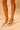 Close-up of a person wearing AZALEA WANG ESMARIA WHITE METAL EMBELLISHED PUMP stilettos with ankle straps and gold accents. Only the lower legs and feet in the faux patent leather heels are visible on a light surface.
