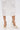 A person in a white lace skirt stands on a white surface, wearing the AZALEA WANG ESMARIA White Metal Embellished Pumps with pointed toes and stiletto heels. Only their lower legs and feet are visible.