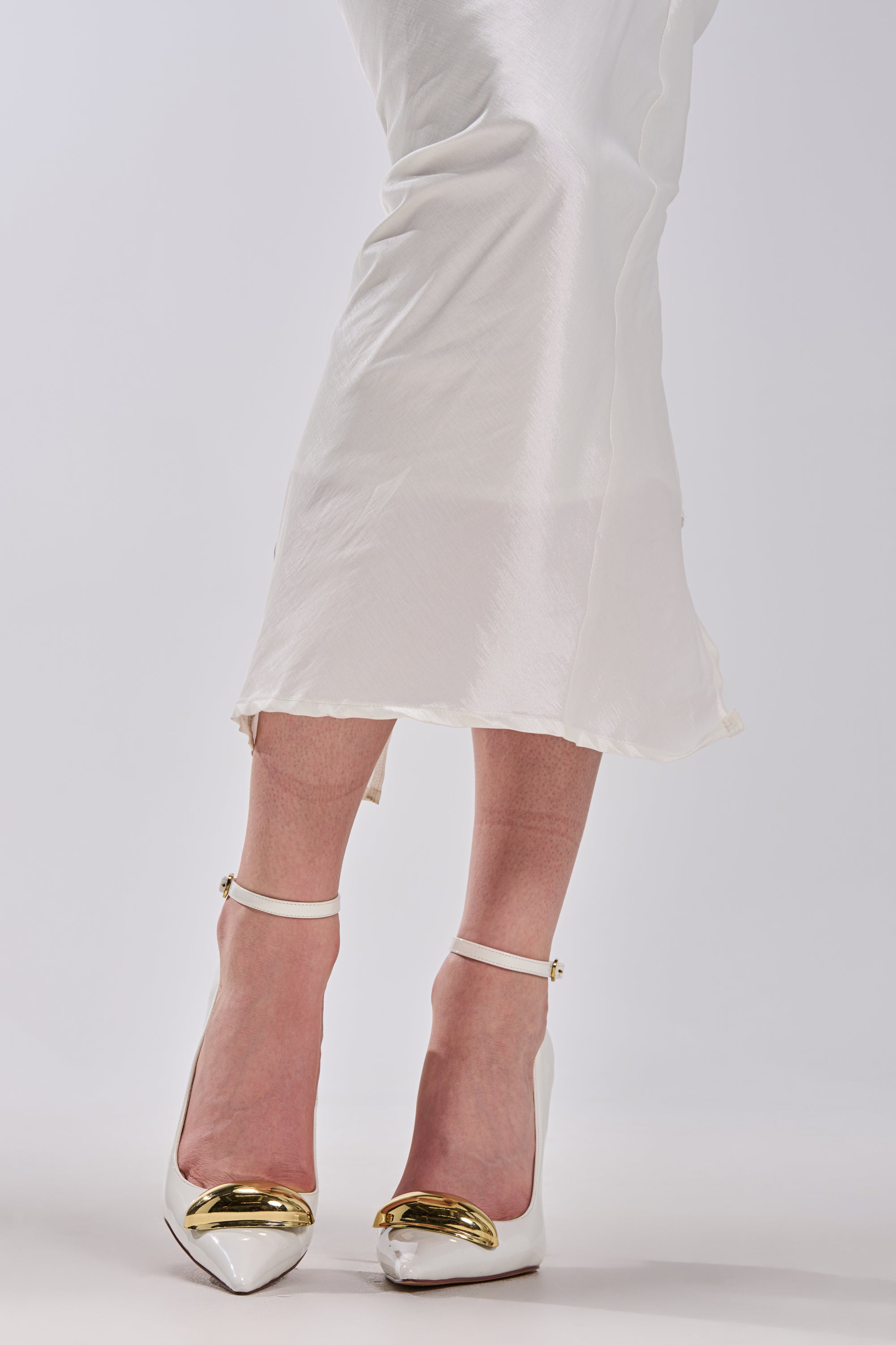 Wearing a long white satin skirt, a person stands with feet crossed against a plain background in AZALEA WANG ESMARIA WHITE METAL EMBELLISHED PUMPS featuring gold detailing and stiletto heels.