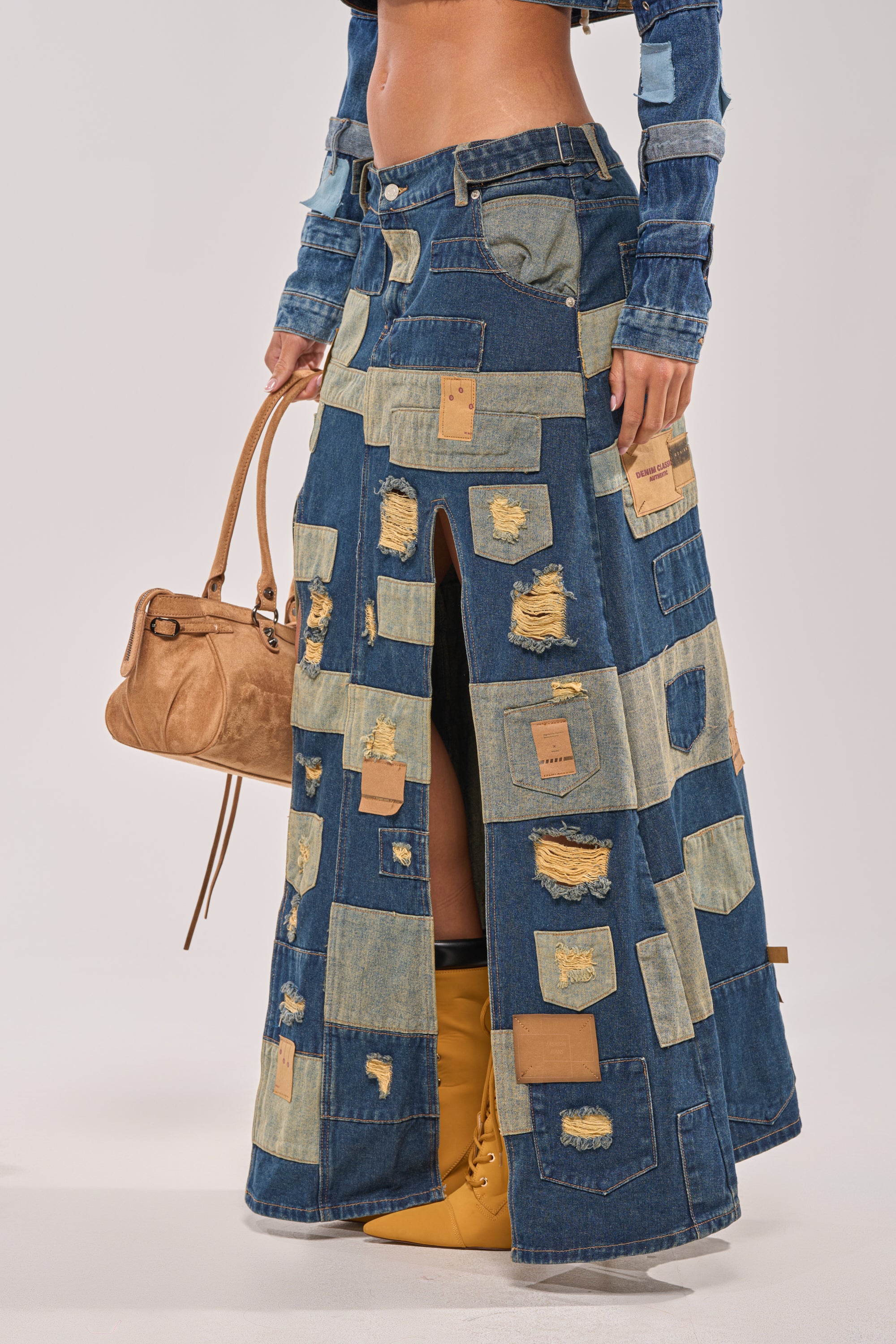 Wearing the PATCH ME UP DENIM MAXI SKIRT with distressed, patchwork details, styled with a brown handbag, cropped denim top, and tan boots—ideal for bold festival streetwear looks. 2