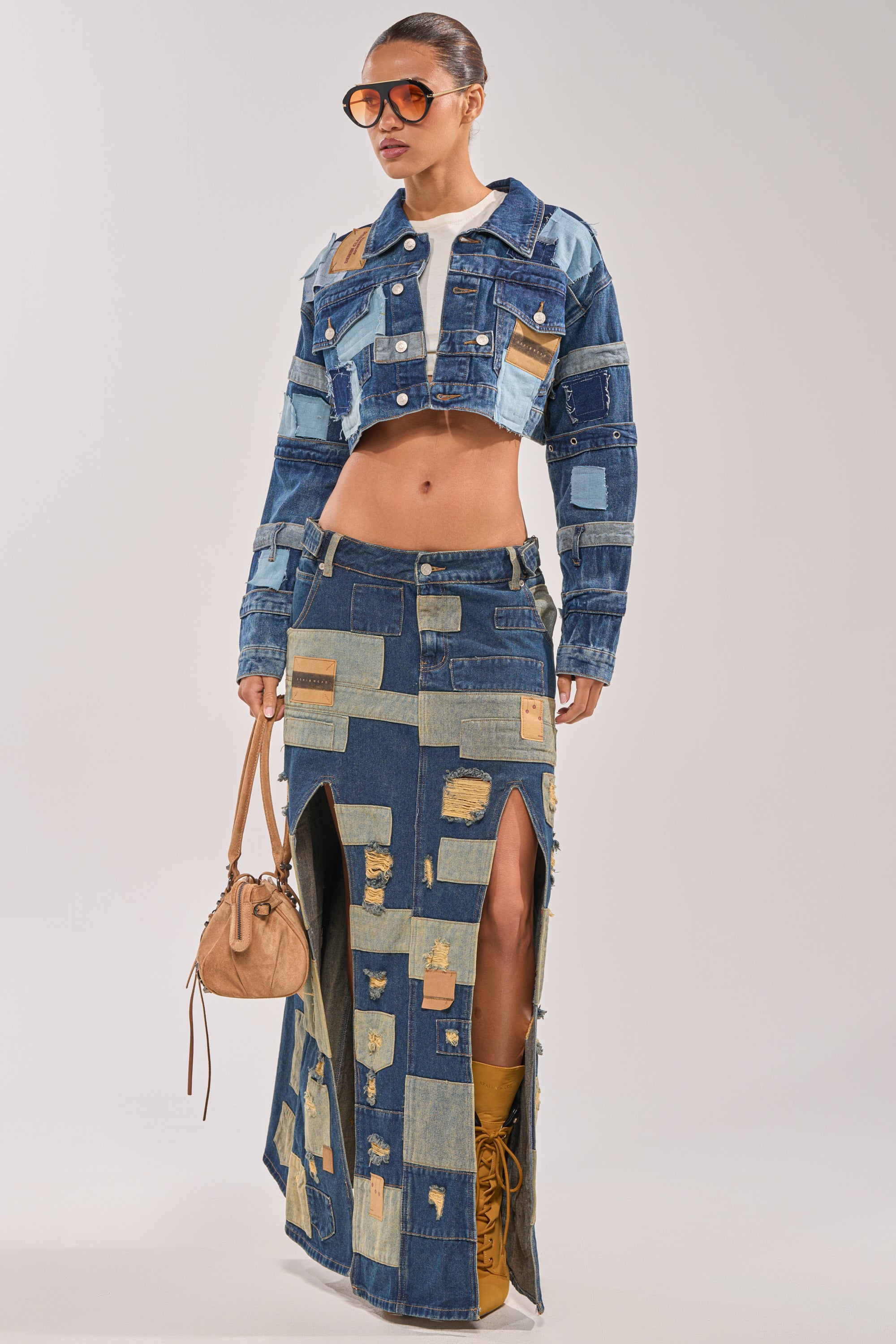 A woman showcases streetwear festival style in the PATCH ME UP DENIM MAXI SKIRT, paired with a matching jacket, yellow boots, sunglasses, and a tan handbag against a plain background. 5