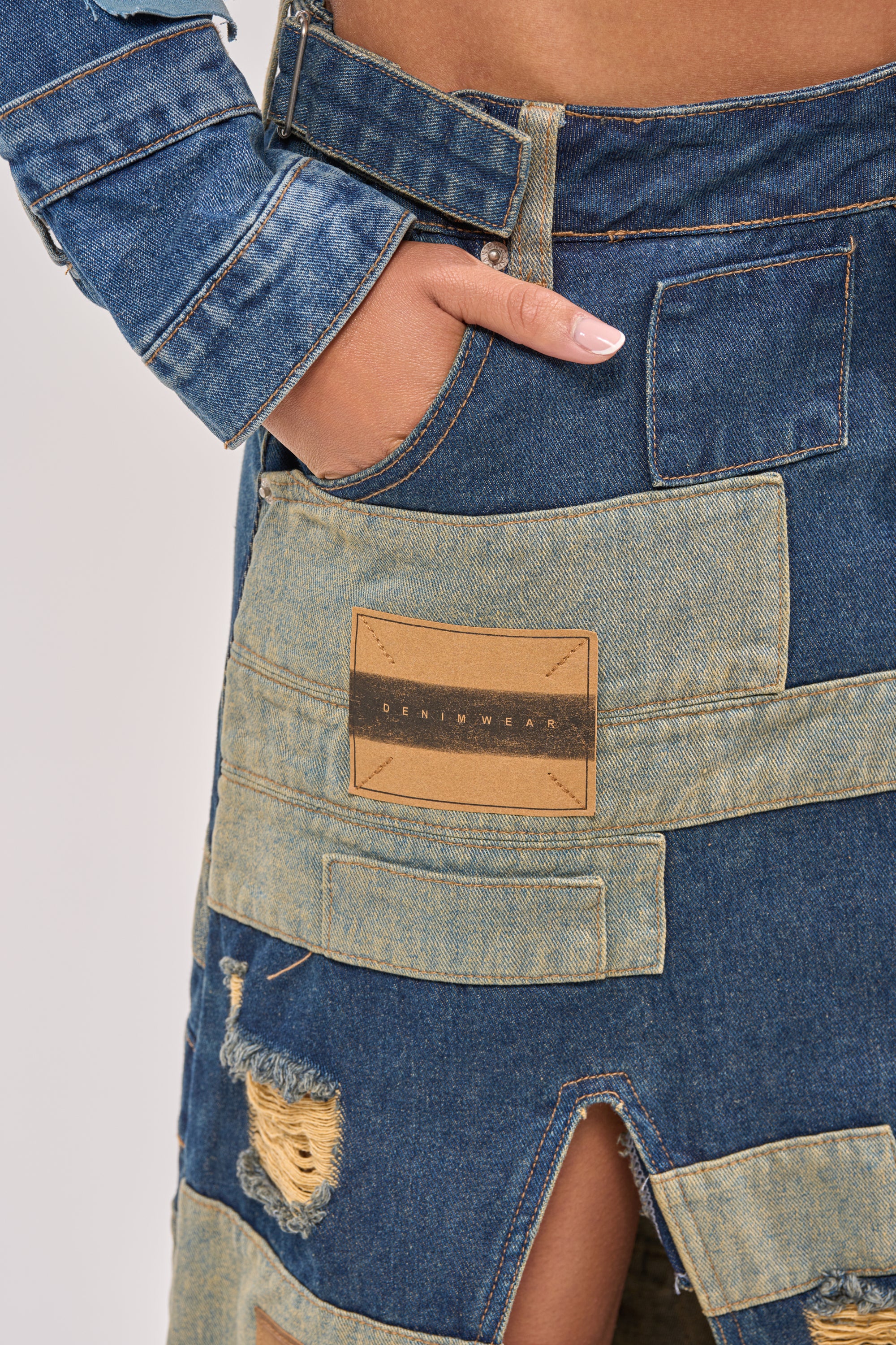 Close-up of a person wearing the PATCH ME UP DENIM MAXI SKIRT, featuring blue patchwork, a front slit, distressed accents, and a "DENIMWEAR" label—ideal for festival vibes or elevating your streetwear style. One hand is tucked in the pocket. 6