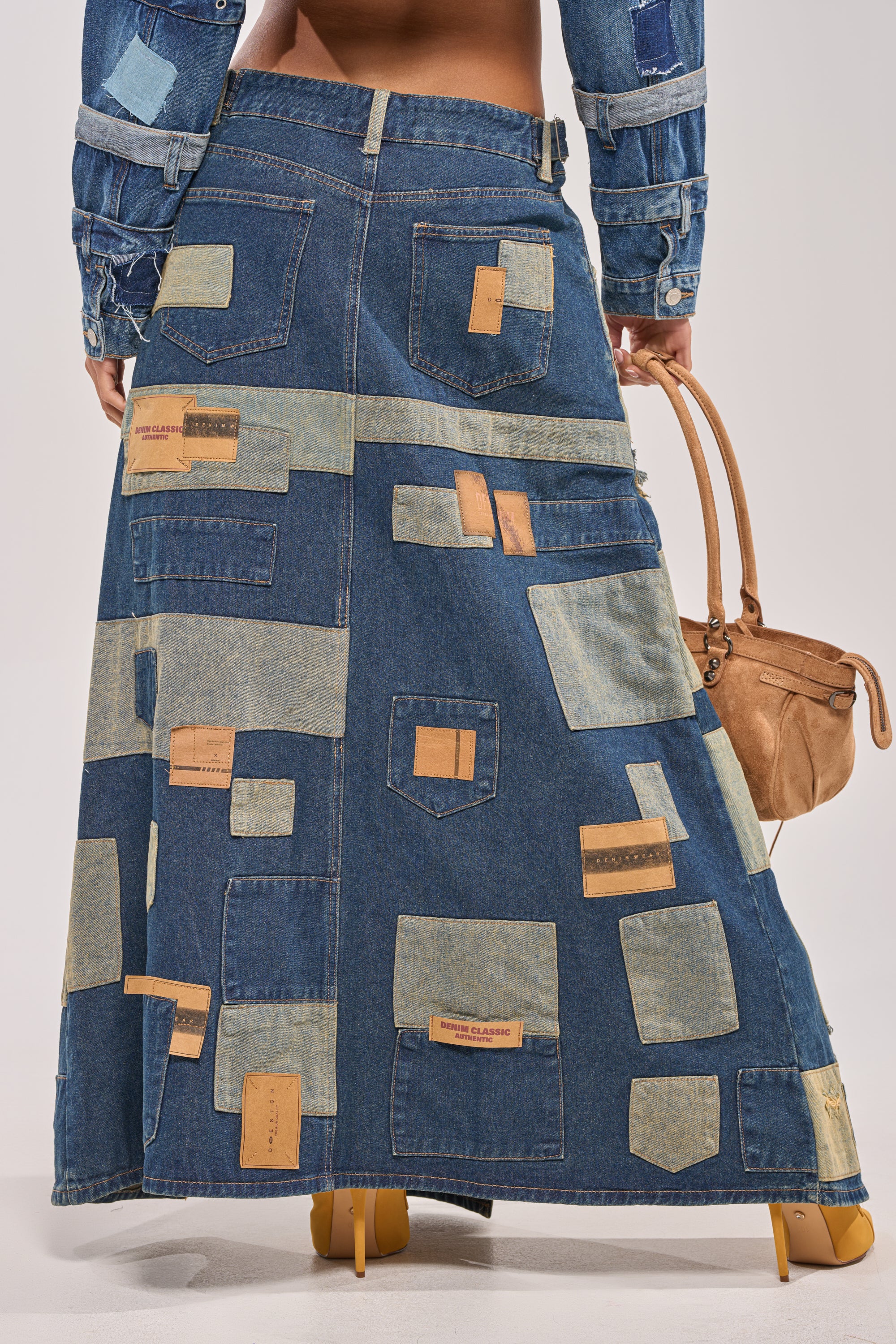 A person is seen from behind in the PATCH ME UP DENIM MAXI SKIRT, featuring patchwork details and pockets, styled with a denim jacket. They carry a tan handbag and wear yellow-heeled shoes—perfect for standout streetwear or festival looks. 4