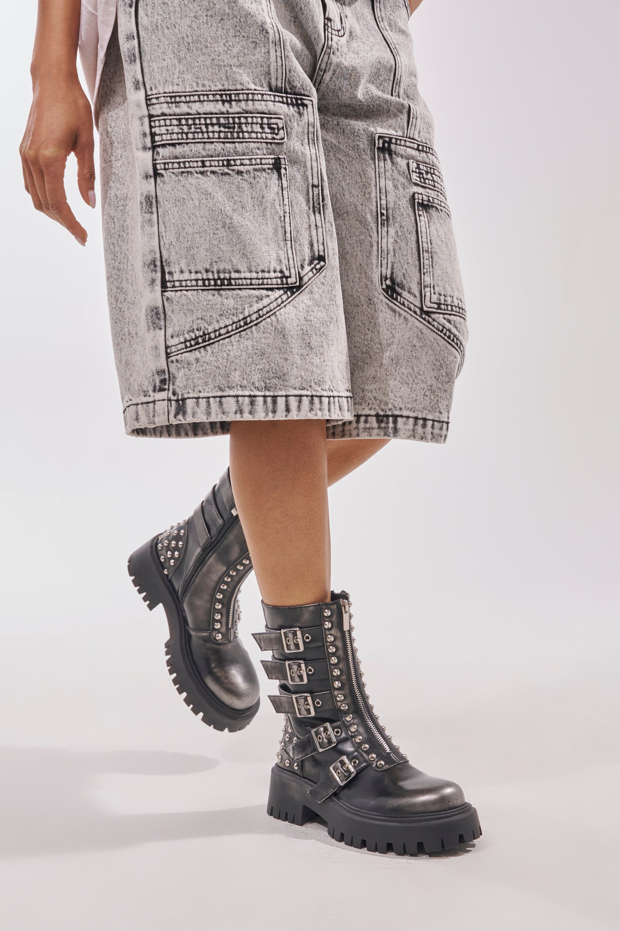 Model wears light-wash denim shorts with large pockets and the AKIRA GOLD ESTERA CHARCOAL EMBELLISHED COMBAT BOOTIE, featuring a chunky heel, multiple buckles, and silver studs, against a plain light background.