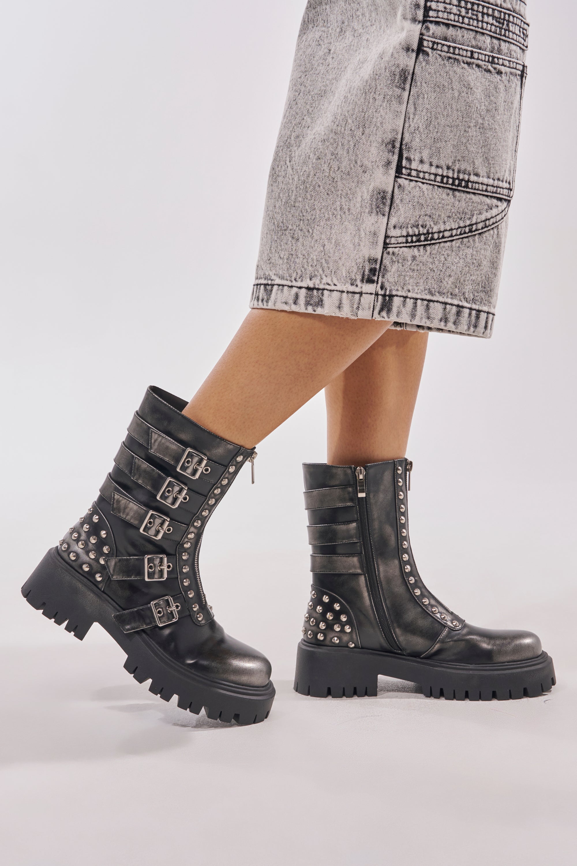 A person in a faded denim skirt and AKIRA GOLD ESTERA CHARCOAL EMBELLISHED COMBAT BOOTIE, standing against a plain light background.