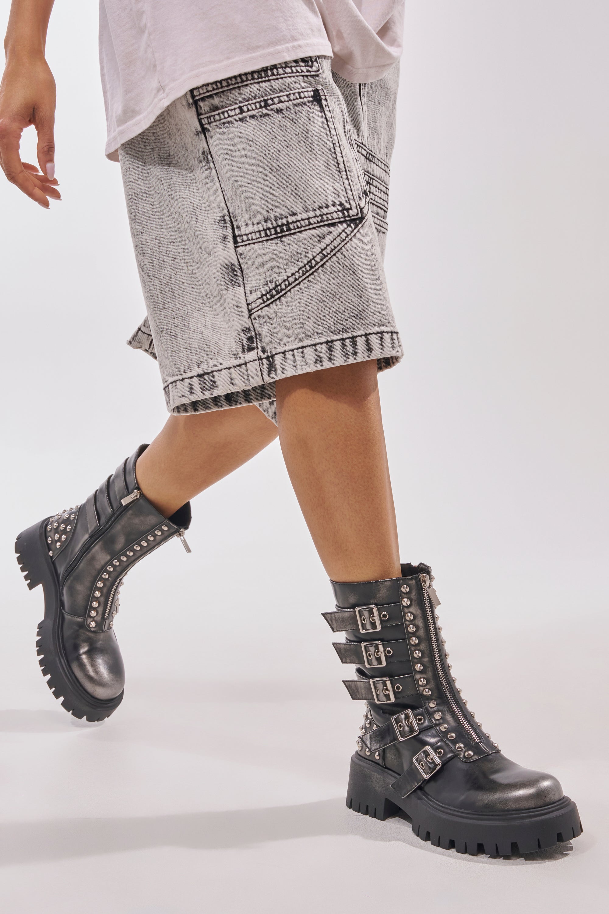 Wearing light acid-wash denim shorts and the AKIRA GOLD ESTERA CHARCOAL EMBELLISHED COMBAT BOOTIE with studs, laces, and multiple buckle straps, a person walks forward against a plain light background.