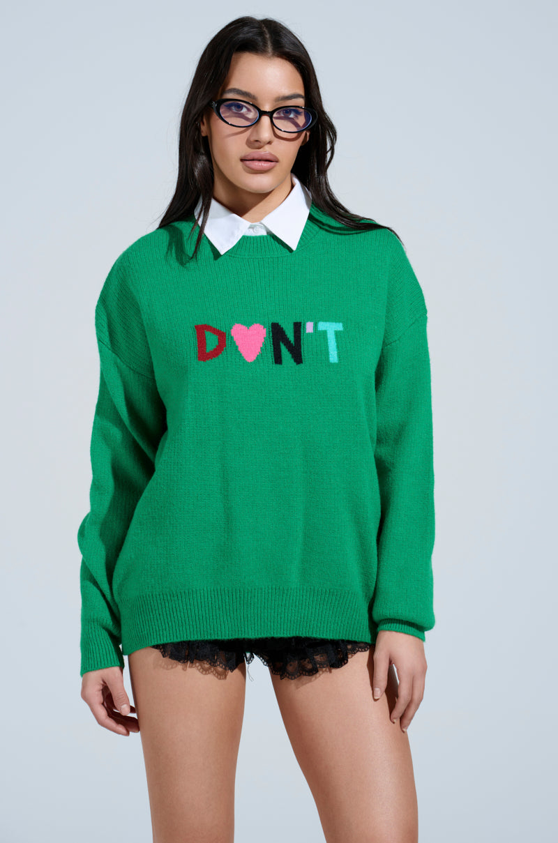A person with long dark hair and glasses wears a green DONT DO IT SWEATER featuring a white collar. Paired with black lace shorts, this bold ensemble sports the design "D♥N'T" across the front and is both daring and unique.