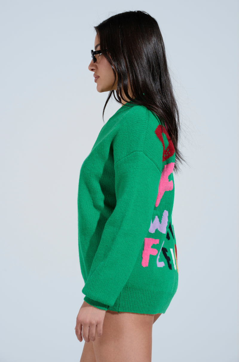 A person with long hair wears sunglasses and the DONT DO IT SWEATER, an oversized knit pullover with colorful abstract letters on the back. They stand sideways against a plain background, exuding style with their cheeky graphic sweater.