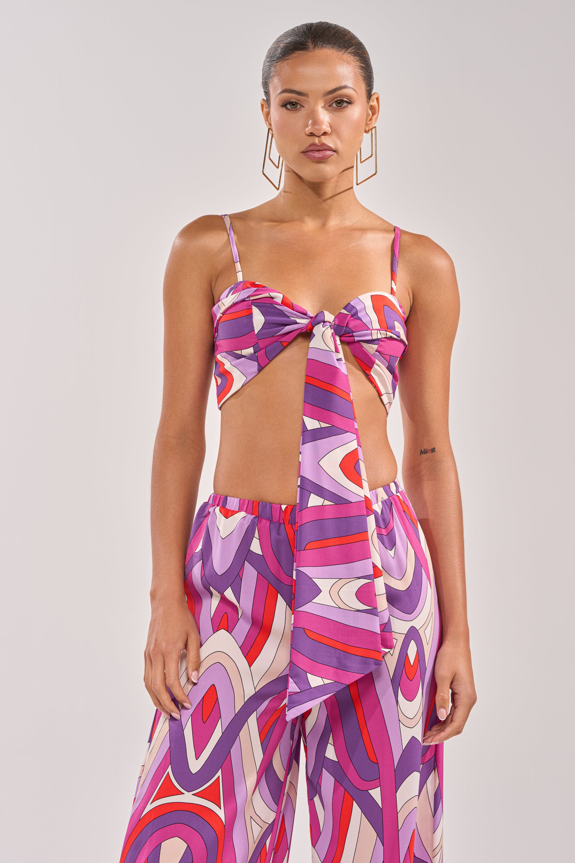 A woman wears the GIRLY POP BRALETTE, featuring vibrant purple geometric patterns and a front tie, paired with matching high-waisted pants for a bold summer look against a light gray background. 1