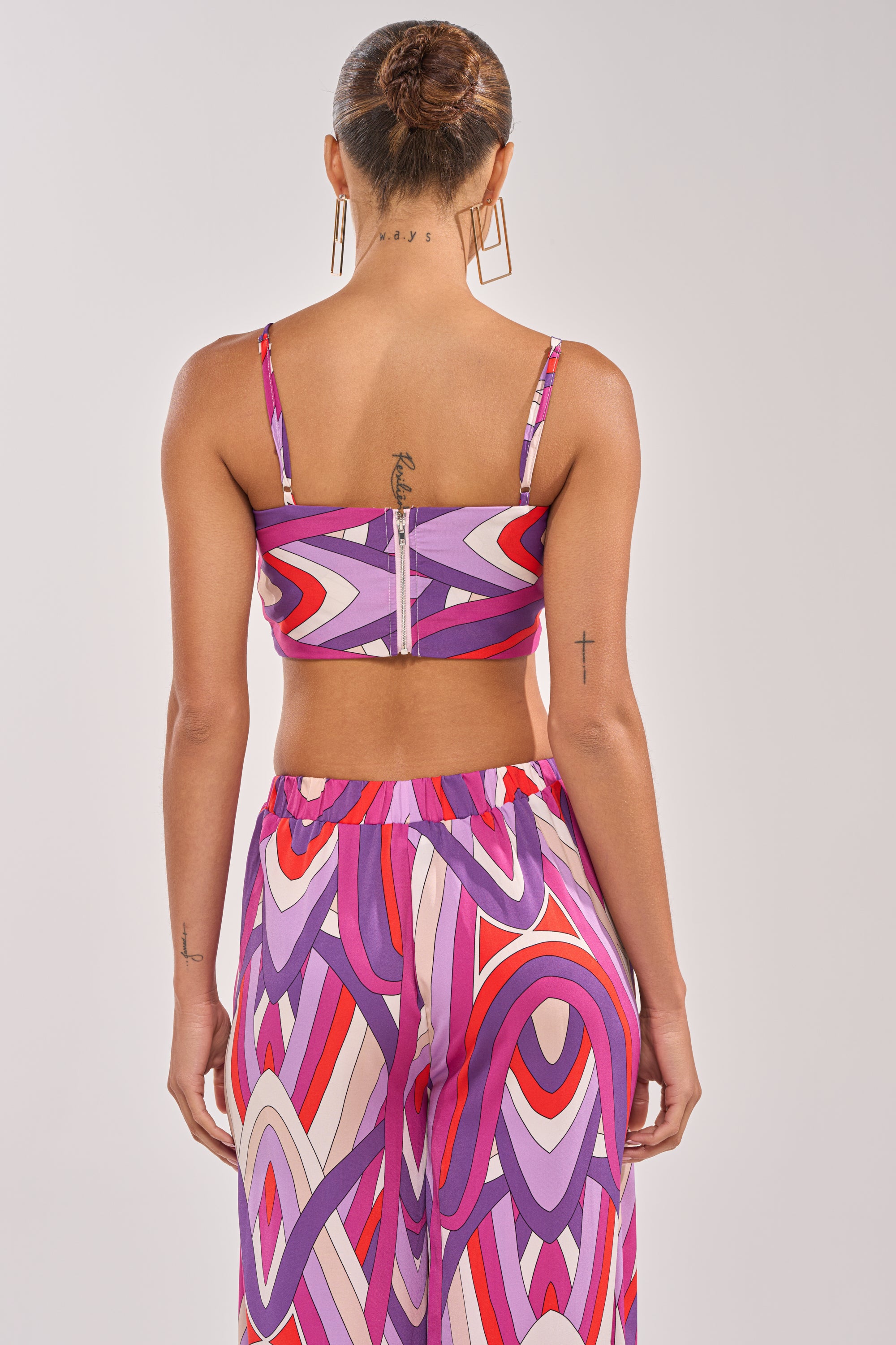 A person stands facing away, wearing the GIRLY POP BRALETTE—a vibrant summer top with thin straps and abstract pink, purple, and red patterns—paired with high-waisted pants. Tattoos and hoop earrings complete the bold look. 3
