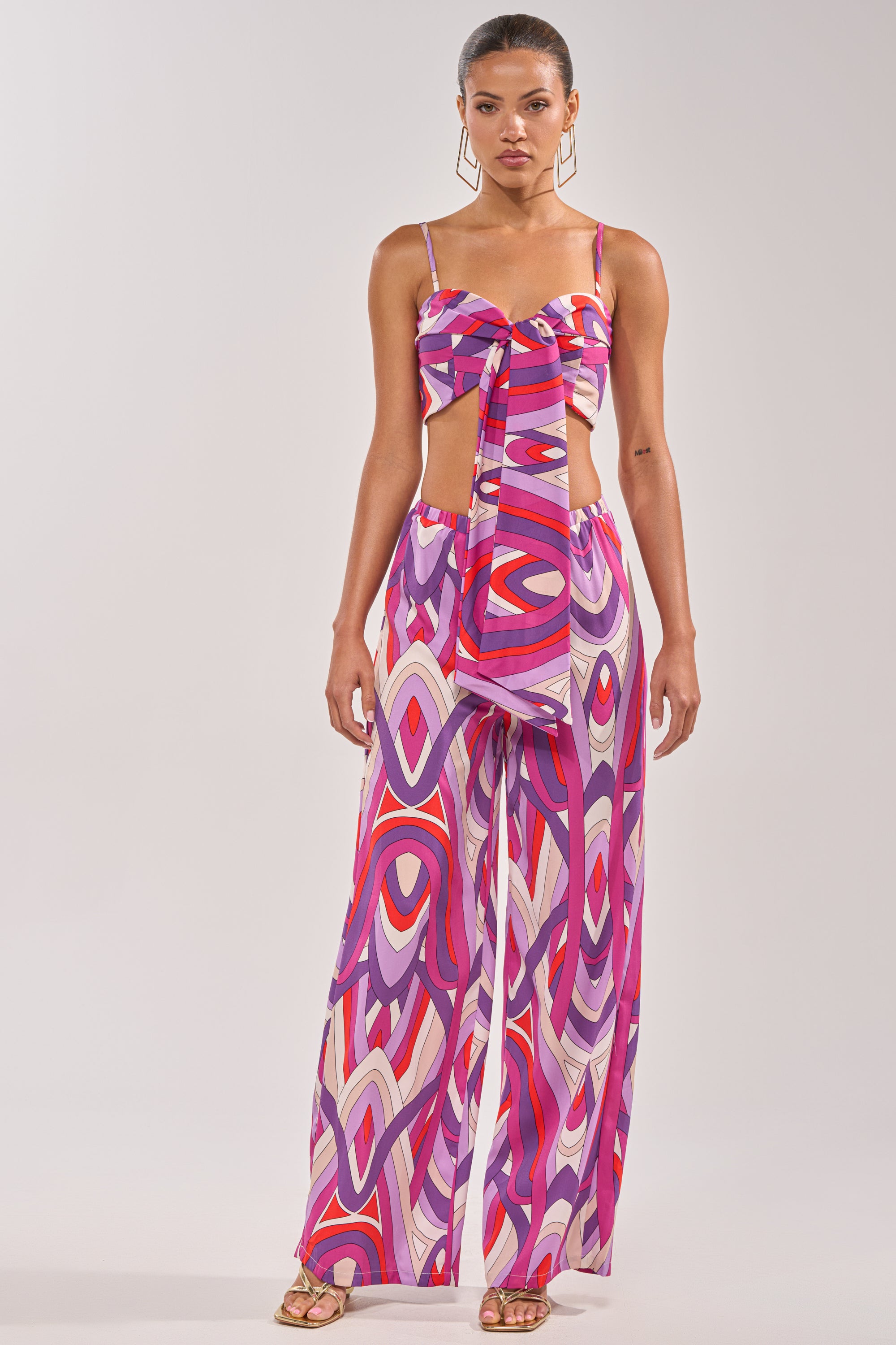 A woman models the GIRLY POP BRALETTE, a colorful geometric-patterned bralette with a front tie, paired with high-waisted wide-leg pants in pink, purple, red, and white. She stands in heels against a plain background. 5