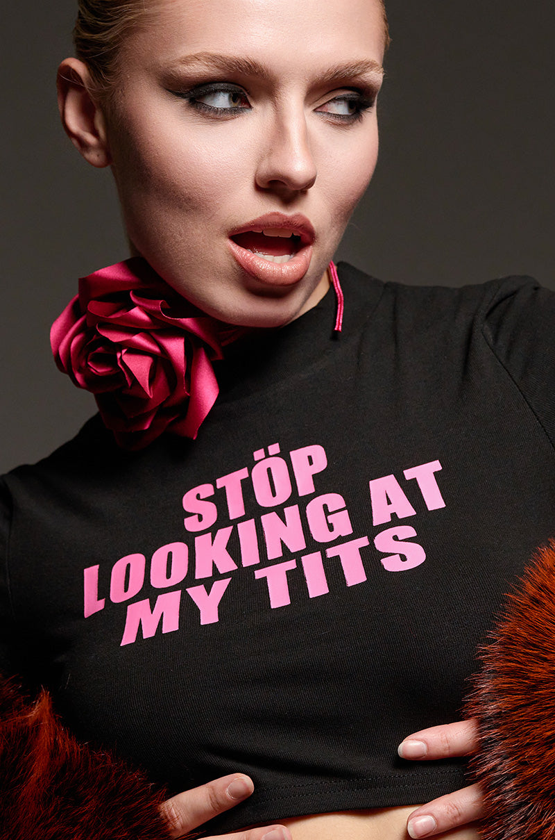 A woman highlights a striking black "STOP LOOKING BABY TEE" featuring bold pink lettering. She complements the look with a large pink flower necklace and sports fur-like red cuffs, all while maintaining a playful expression with her mouth slightly open.