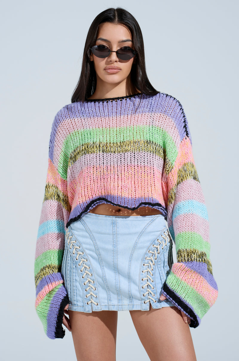 A person confidently poses in the SURE THANG SWEATER, featuring a chunky striped pattern. With long hair, they sport an oversized look paired with a light blue denim skirt with lace-up sides. The plain, light blue background is ideal for Spring sweater season.