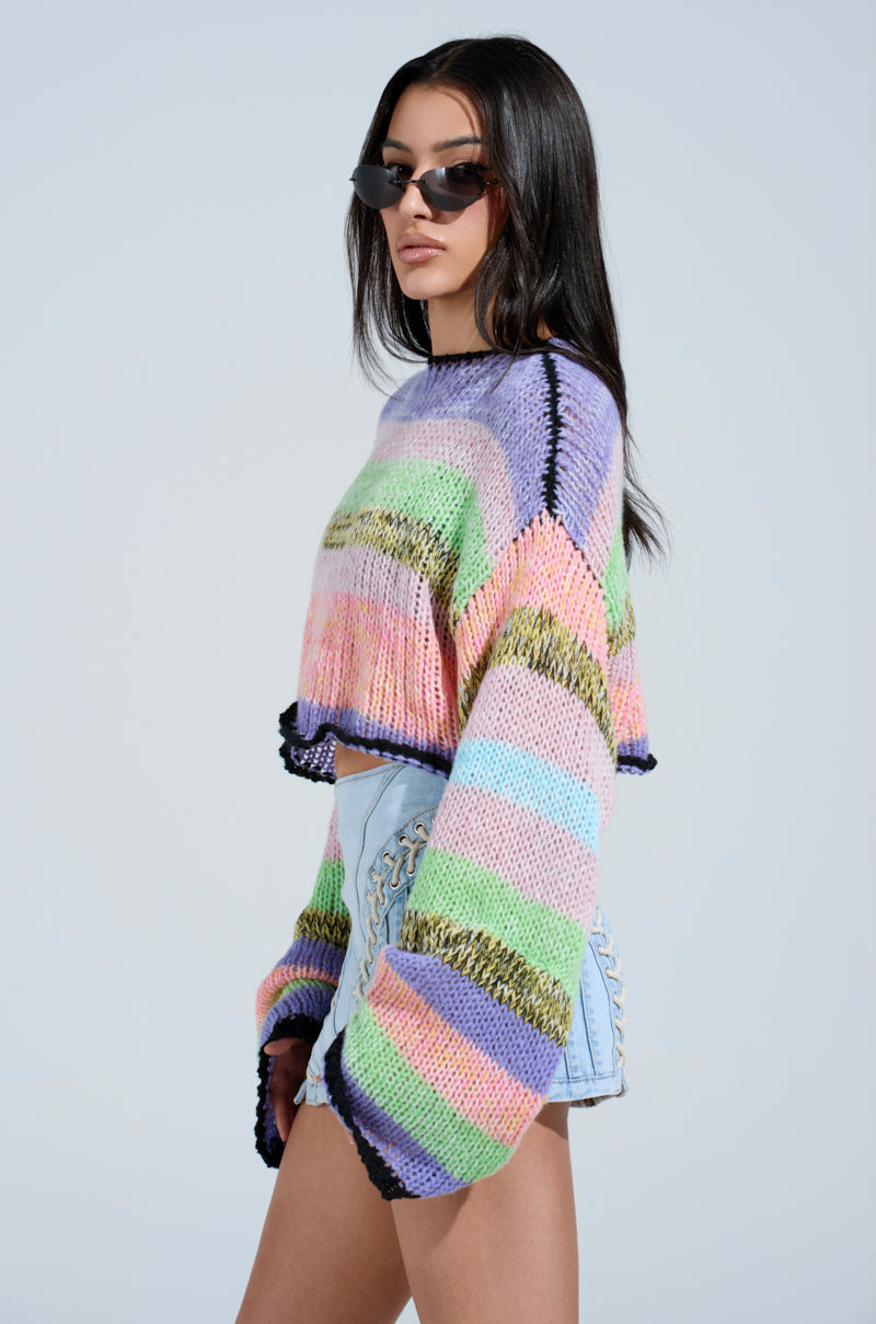 Sporting long hair and sunglasses, they wear the SURE THANG SWEATER, a colorful chunky knit with stripes, paired with light blue denim shorts. The oversized silhouette enhances their style as they pose against a plain background.