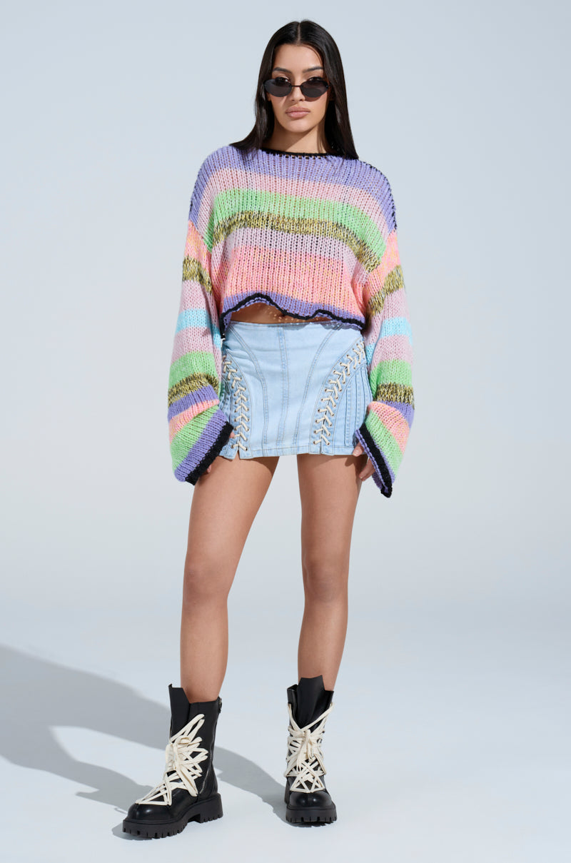 A person wearing the SURE THANG SWEATER, a light blue denim skirt with lace-up details, and black boots with white laces. They also have dark sunglasses on while standing against a plain background.