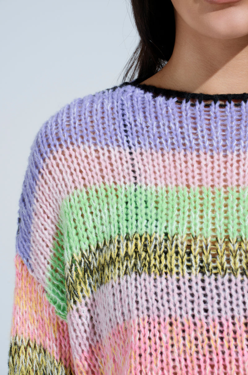 A close-up shows someone wearing the SURE THANG SWEATER, an oversized knit with chunky stripes in purple, pink, green, yellow, and black. The textured stitch enhances the cozy vibe while their face peeks against a light background, celebrating spring sweater joy.