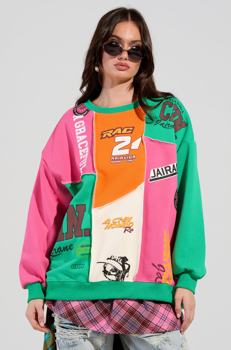A woman wearing sunglasses and confidently sporting an AKIRA Label MIXED EMOTIONS SWEATSHIRT, boldly adorned with vivid logos and text, stands out. The sweatshirt features a striking color block pattern in shades of pink, green, and orange, which she expertly pairs with a plaid skirt accented by frayed edges. Her stylish ensemble is highlighted against the plain background.