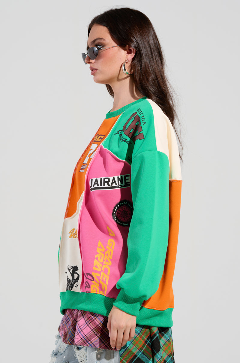 A woman with long dark hair, sporting sunglasses and hoop earrings, is wearing the vibrant MIXED EMOTIONS SWEATSHIRT adorned with racing-inspired graphics. She pairs it with a plaid skirt featuring a mesh overlay, set against a simple gray backdrop.
