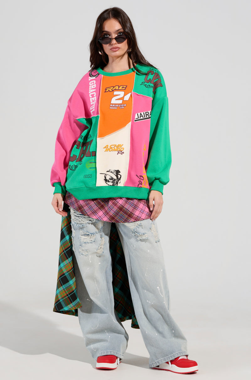 A person wearing a vibrant MIXED EMOTIONS SWEATSHIRT adorned with racing-inspired graphics, styled over a pink plaid shirt. They pair it with light blue distressed jeans and red sneakers. The outfit is completed with black sunglasses and features a bold touch from the plaid skirt accent.