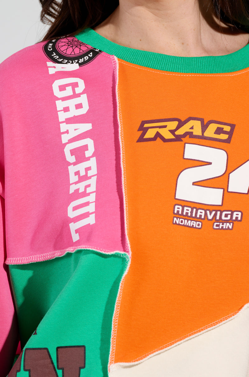 A close-up of someone wearing the MIXED EMOTIONS SWEATSHIRT, which boasts racing-inspired graphics in a colorful patchwork design. The sweatshirt features vivid sections of pink, orange, green, and cream, adorned with printed text and images such as "GRACEFUL," the number "24," and other vibrant color block patterns.