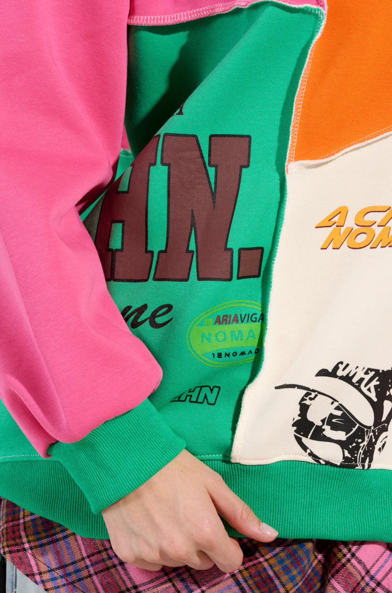 A person wearing a colorful MIXED EMOTIONS SWEATSHIRT and a plaid skirt holds the edge of the sweatshirt, showcasing its vibrant, racing-inspired graphics and color block pattern that combine logos and text in green, pink, orange, and cream.