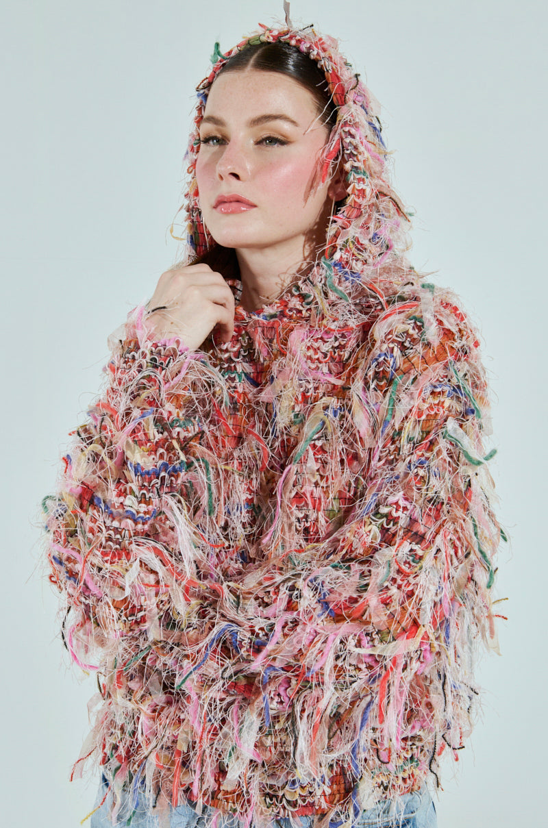 A person wears the ALL OVER THE PLACE FRINGE SWEATSHIRT, featuring a textured design with vibrant multicolored threads creating a shaggy look. The plain, light background highlights the garment's unique and lively fringe trim.