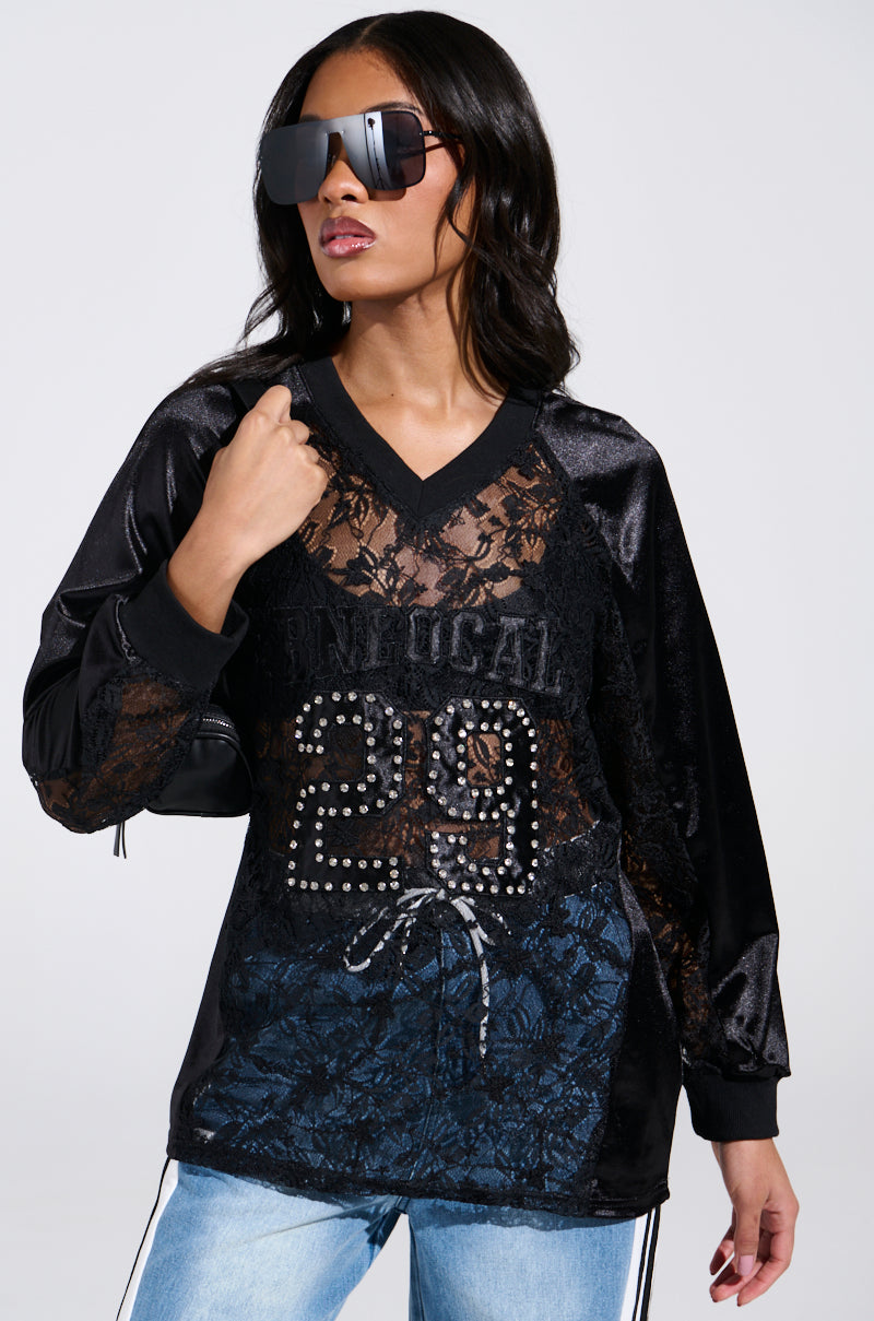 A woman wears the KIKI LACE JERSEY, adorned with "29" and "FIND US ALL" in studded letters. Her long dark hair falls over sporty-chic sunglasses. Completed with light blue jeans, she effortlessly holds a bag strap over her shoulder, exuding confidence and style.