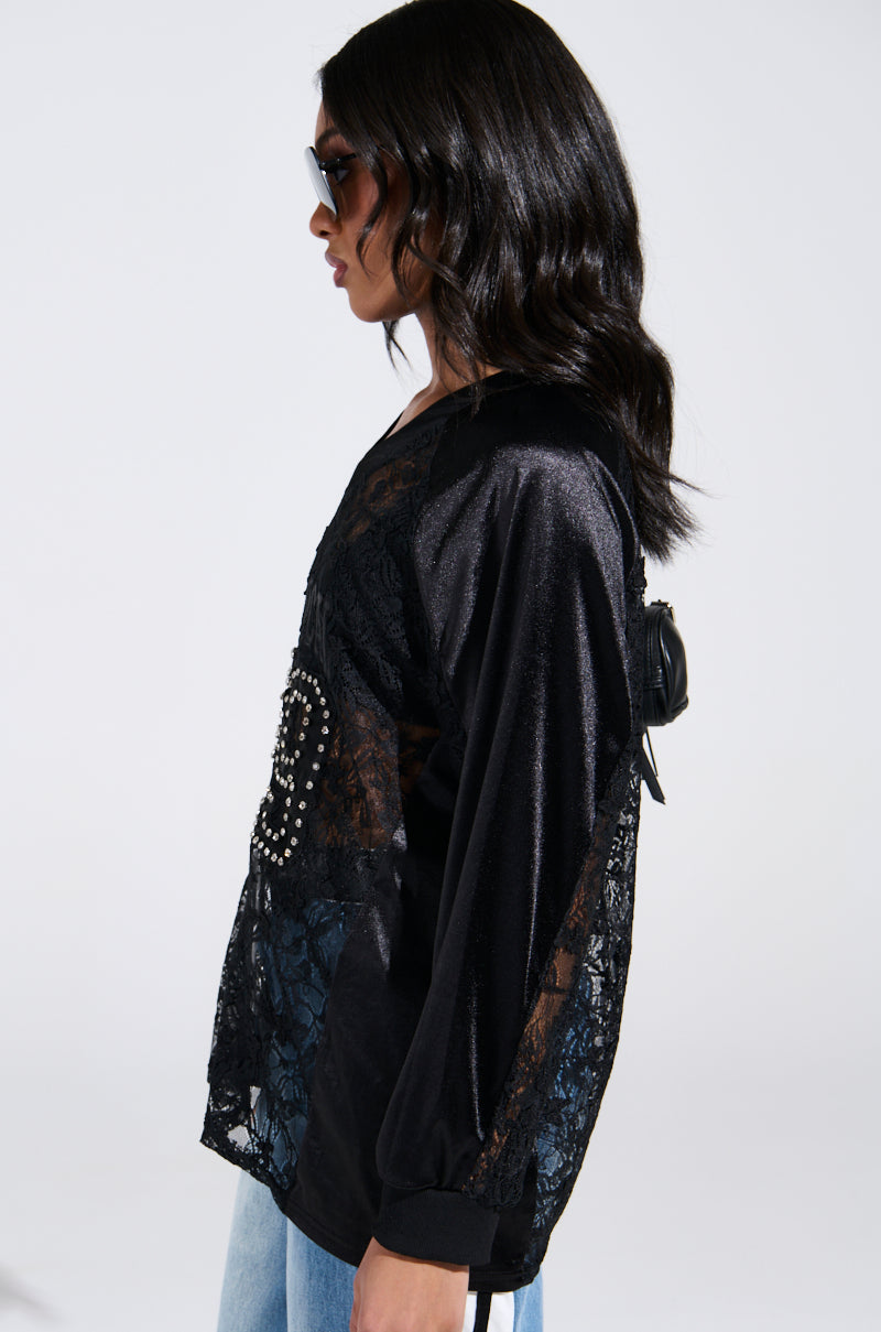 A woman with long dark hair and sunglasses wears the KIKI LACE JERSEY, featuring studded details, matched with light blue jeans. She stands sideways against a plain background, embodying an edgy yet sporty-chic style.