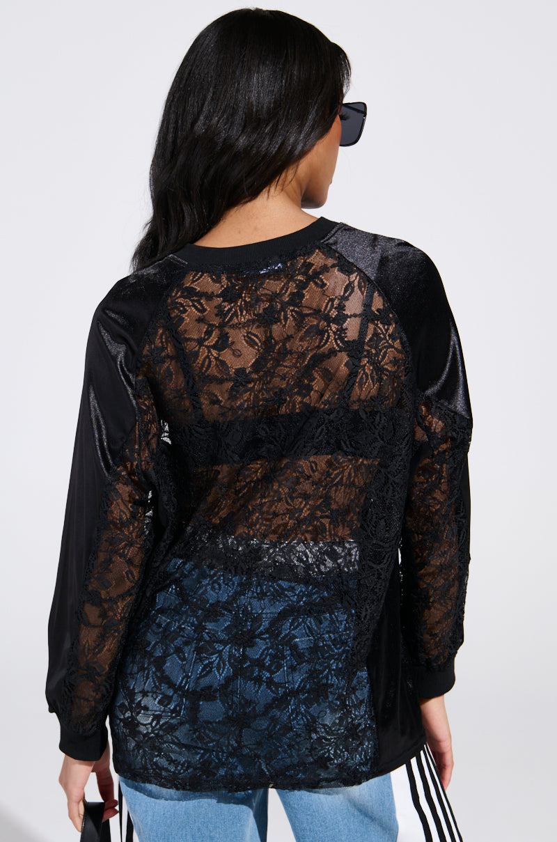 A person with long dark hair is facing away, showcasing a sporty-chic style in sunglasses and the KIKI LACE JERSEY, a black long-sleeve top with sheer floral lace panels on the back, paired perfectly with blue jeans featuring white side stripes.
