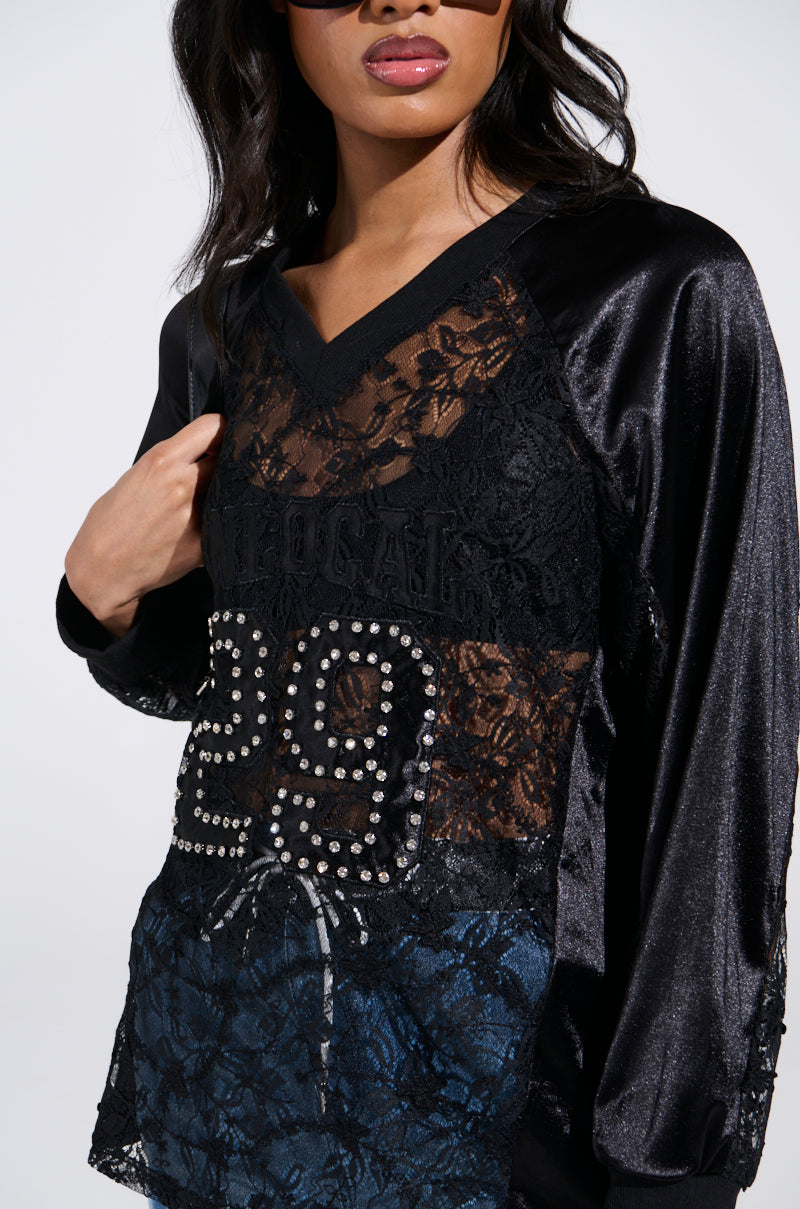 A woman wearing black sunglasses confidently flaunts the KIKI LACE JERSEY, featuring a sporty-chic design with a floral lace bodice and satin textures. An embellished "29" adorns the front for a bold statement.