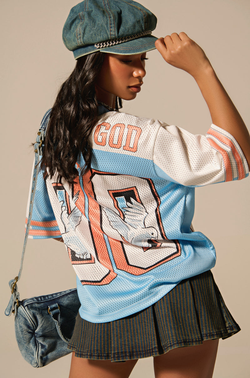 A person models the FLY AWAY JERSEY, featuring sporty accent stripes and "GOD 00" adorned on the back. The color-block ensemble includes a cap, striped skirt, and is paired with a casually slung denim bag.