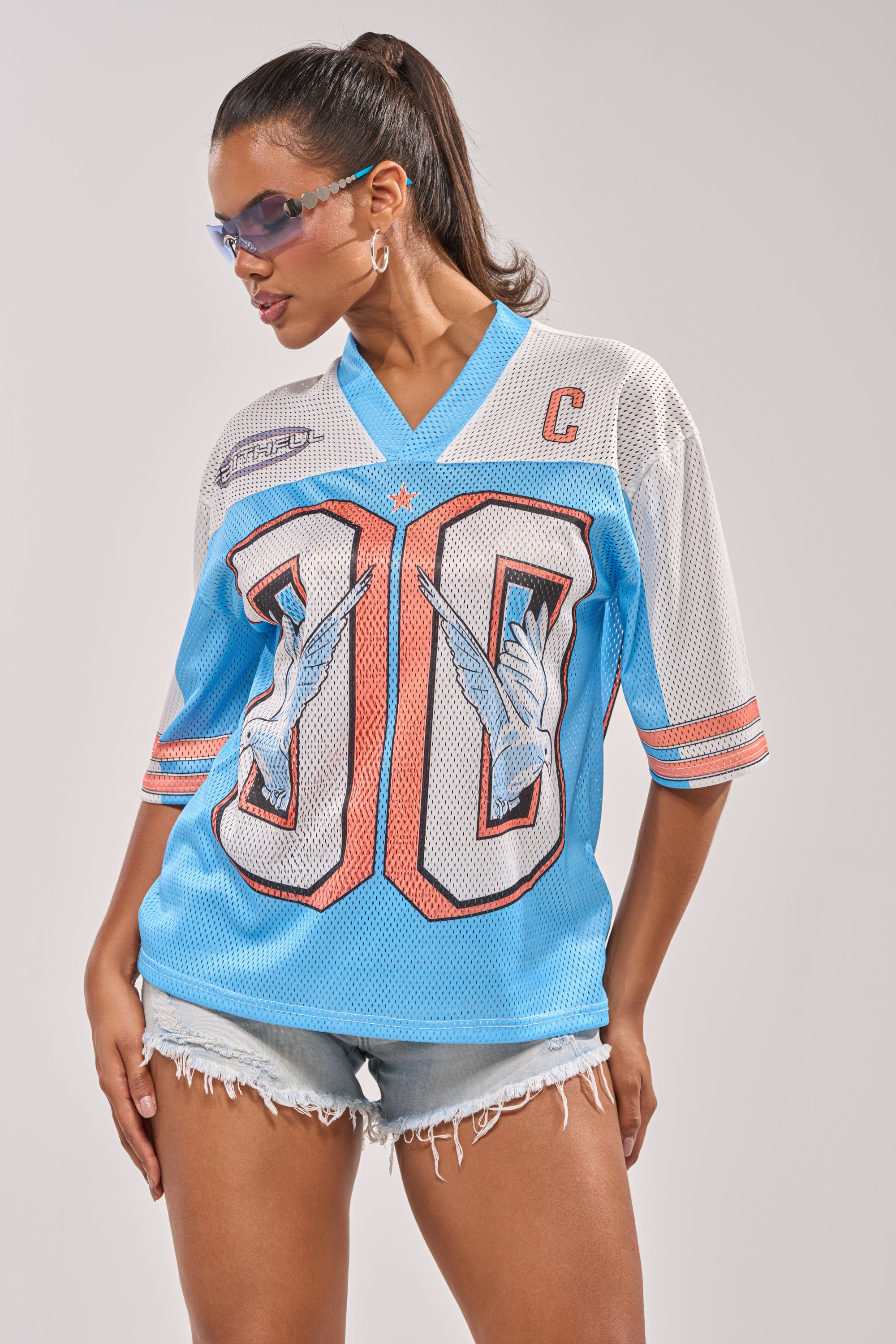 A woman models the FLY AWAY JERSEY, featuring bird graphics and the number 90, paired with blue-tinted sunglasses, hoop earrings, and distressed denim shorts for a sporty streetwear look against a plain background. 3