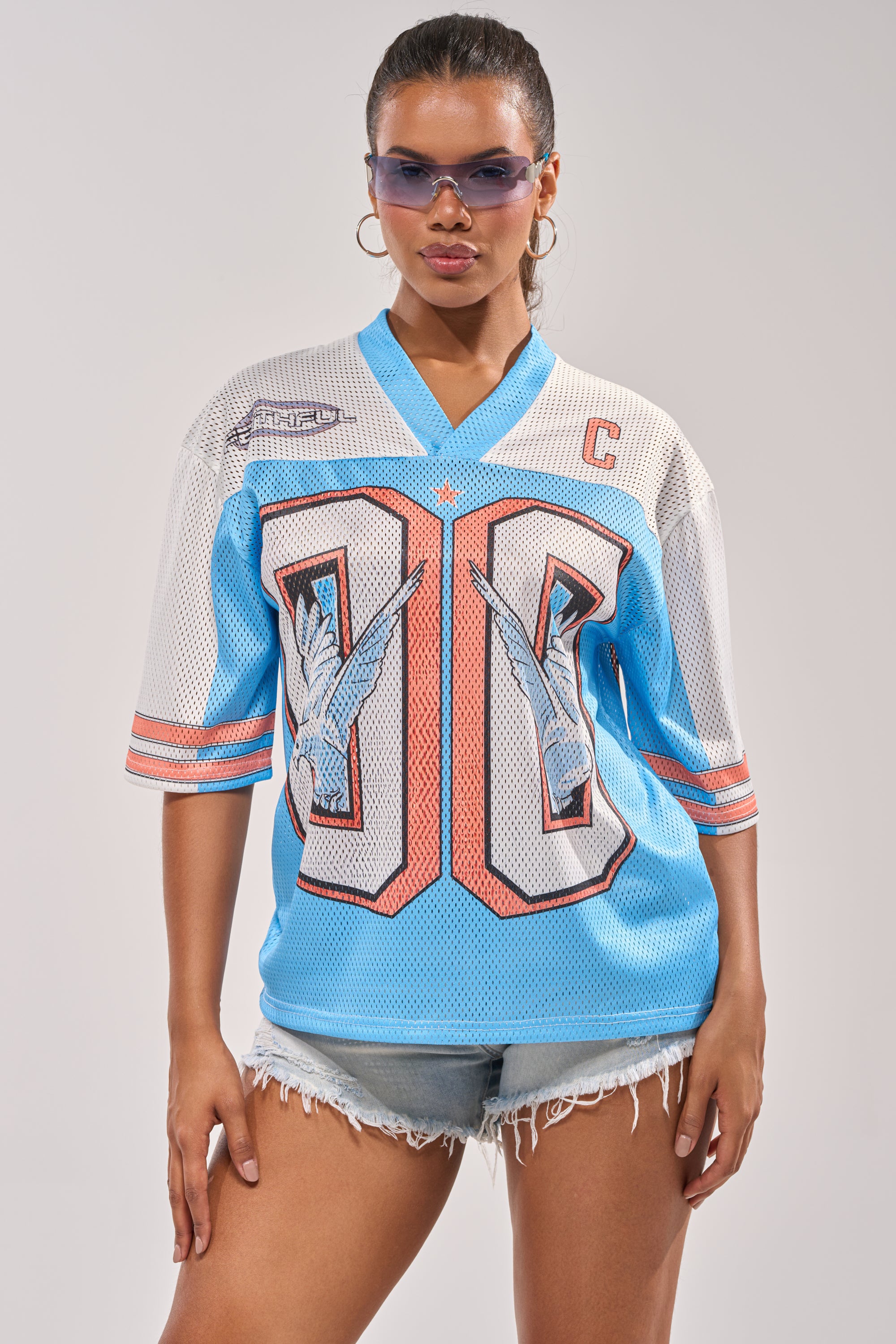 A woman poses confidently against a plain light background, wearing sunglasses, distressed denim shorts, and the FLY AWAY JERSEY—a blue and white varsity top with large "00" and eagle graphics. 4