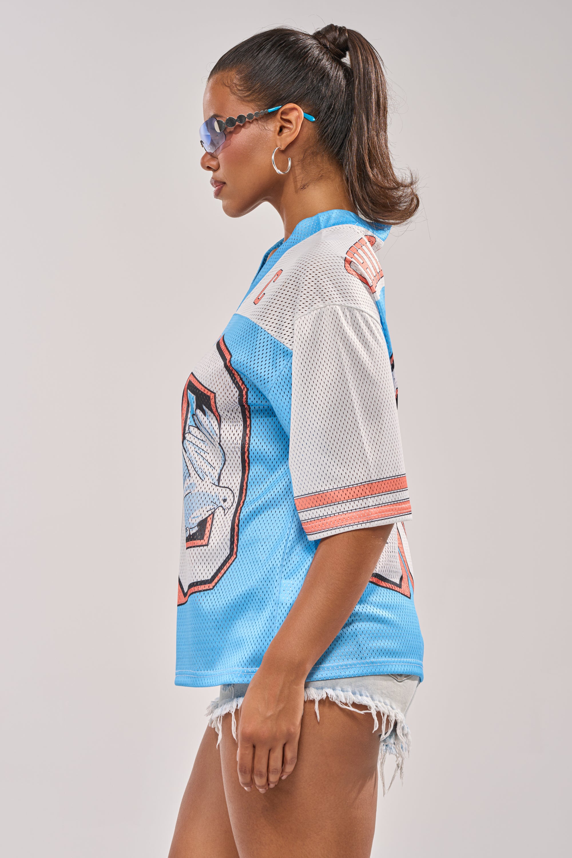 A woman with a ponytail wears sunglasses, frayed denim shorts, and the FLY AWAY JERSEY, standing in profile against a plain background. 5