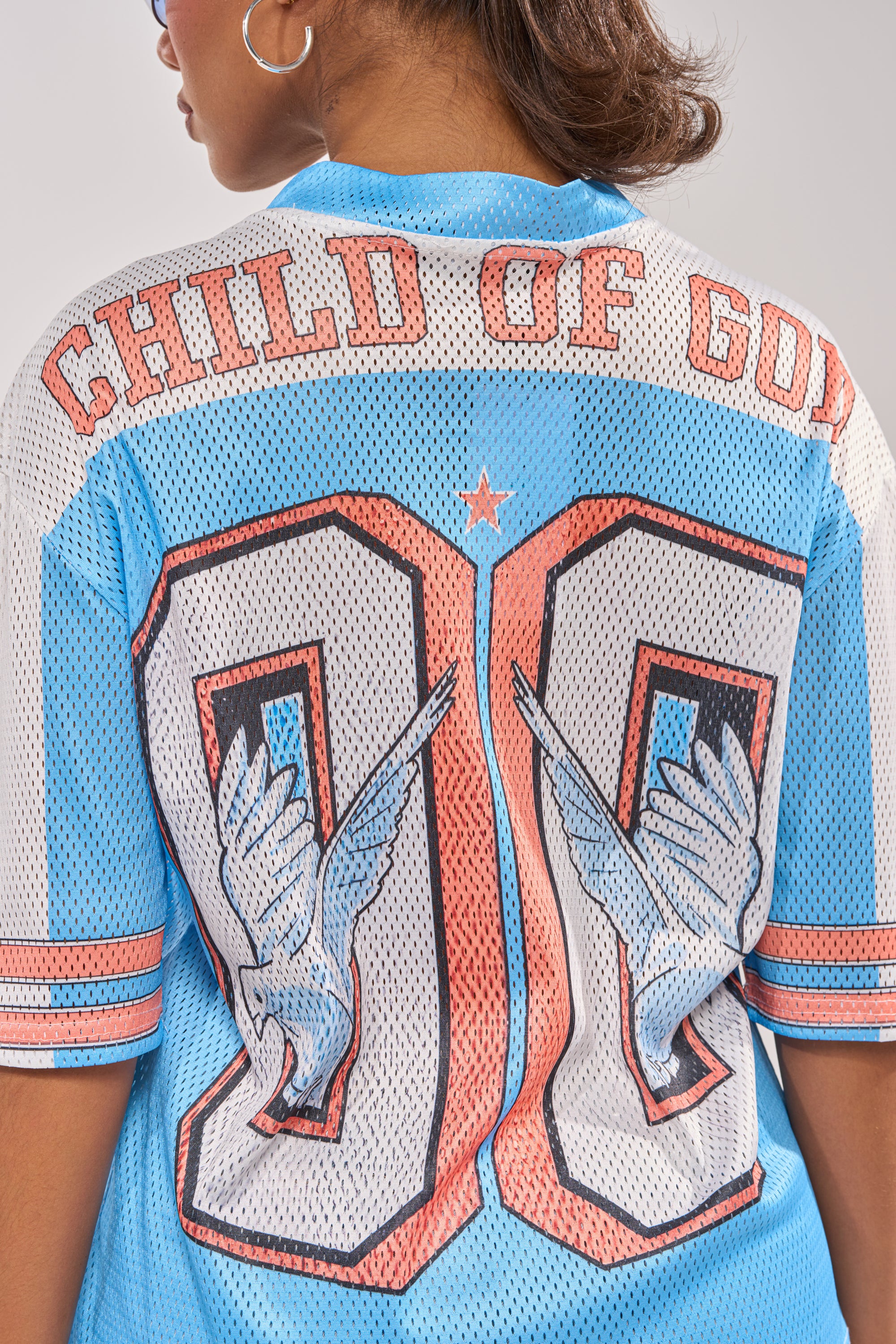 A person wears the FLY AWAY JERSEY, a sporty blue mesh varsity top with "CHILD OF GOD" and bold "99" on the back, accented by white doves—merging classic streetwear vibes with a unique spiritual flair. 9