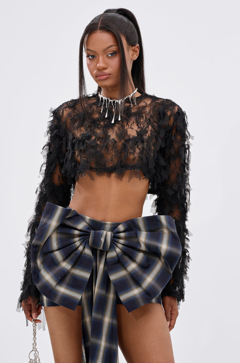 A woman stands confidently in the THOUGHT YOU KNEW CROPPED FRINGE TOP, paired with a large blue plaid bow skirt, a statement necklace, and a chain handbag against a plain background.