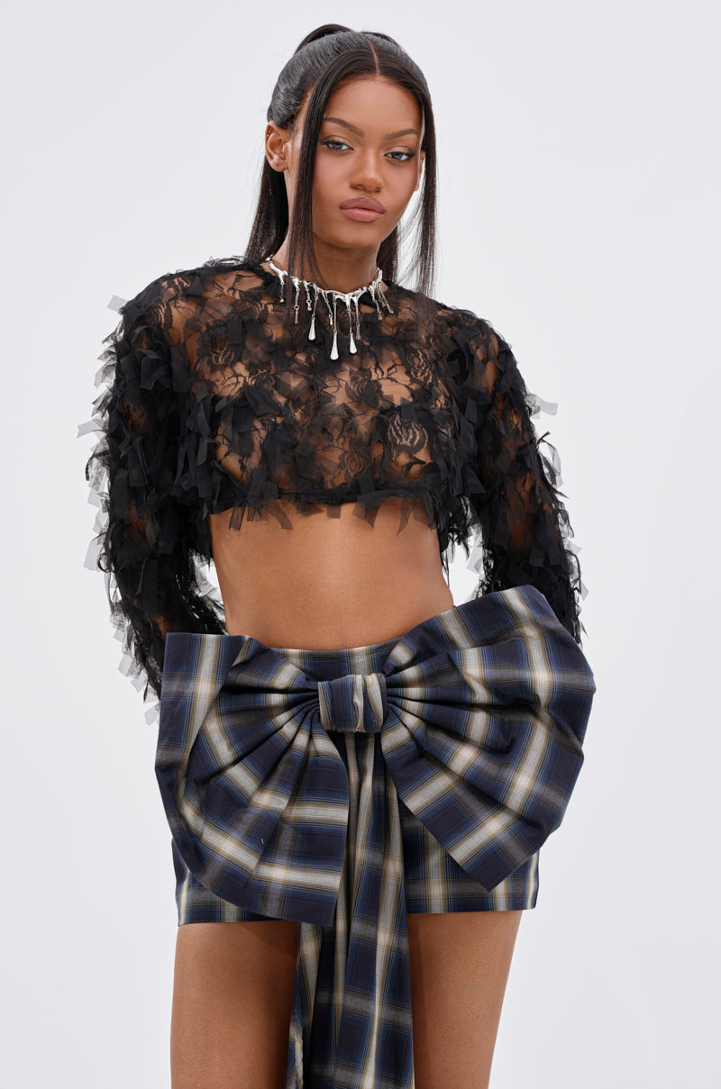 A woman wears the AKIRA Label THOUGHT YOU KNEW CROPPED FRINGE TOP with a large plaid bow skirt. She has long, straight hair, a serious expression, and a silver necklace against a plain, light-colored background.