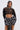 A woman wears the AKIRA Label THOUGHT YOU KNEW CROPPED FRINGE TOP with a large plaid bow skirt. She has long, straight hair, a serious expression, and a silver necklace against a plain, light-colored background.