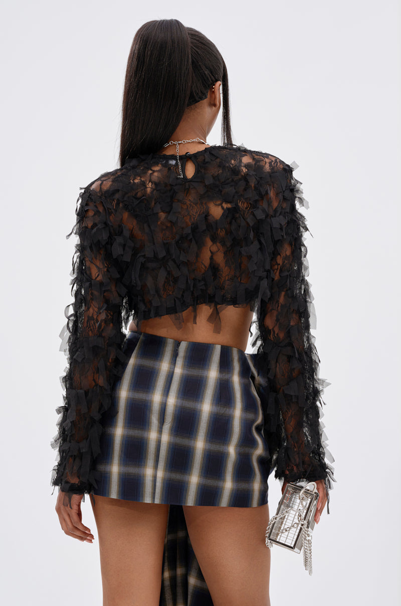A woman with long, dark hair in a high ponytail is seen from behind, wearing the THOUGHT YOU KNEW CROPPED FRINGE TOP and a blue, white, and yellow plaid mini skirt. She holds a small, shiny handbag in her right hand.