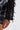 A close-up shows an arm and hand wearing the THOUGHT YOU KNEW CROPPED FRINGE TOP, featuring a blue plaid body and a black, textured, semi-sheer sleeve with layered fabric strips against a plain white background.